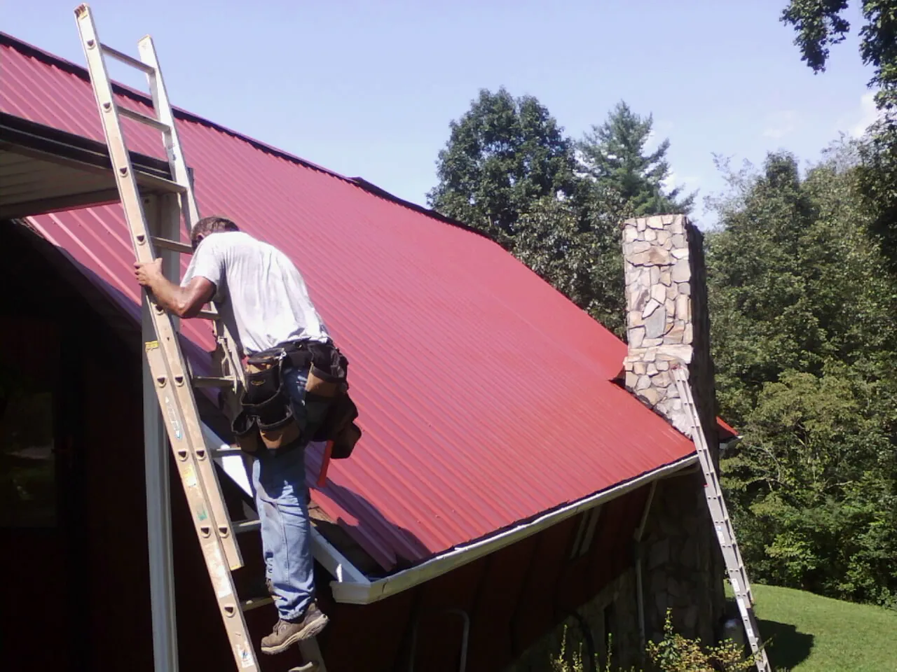 Roofer on a ladder beside a red metal roof and stone chimney — Soffit & Fascia Repair by Roofing Brothers in Port Townsend